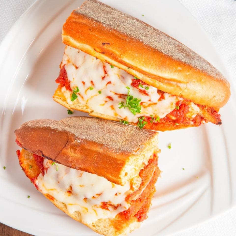 Two halves of a chicken parmesan hero sandwich, topped with melted mozzarella and parsley, on a white plate.